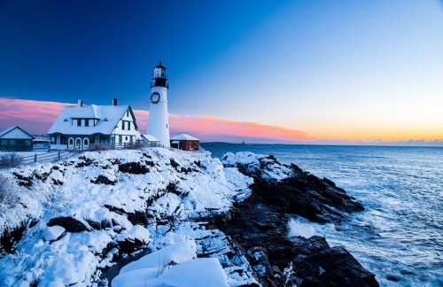 A snowy lighthouse stands on rocky shores at sunset, with a vibrant sky and calm ocean waves in the background.