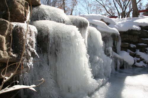A frozen waterfall cascading over rocks, surrounded by snow-covered trees under a clear blue sky.