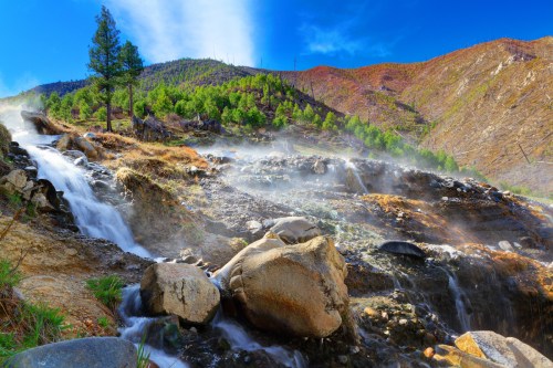 A serene landscape featuring a flowing stream, rocky terrain, and mist rising against a backdrop of green hills and blue sky.