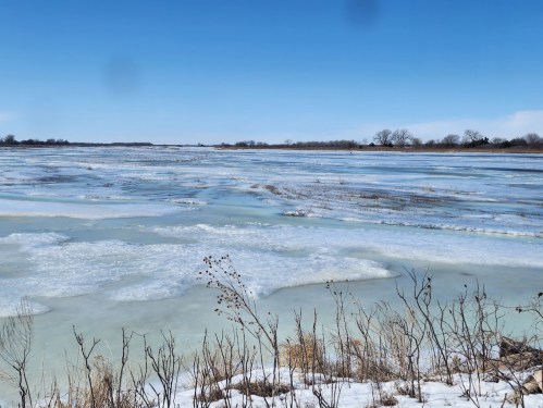A frozen river landscape under a clear blue sky, with patches of ice and snow along the banks.