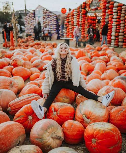 A smiling woman sits among large pumpkins at a festive outdoor pumpkin patch.