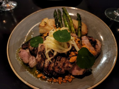 A plate of grilled meat with sliced apples, nuts, asparagus, and potatoes, garnished with green leaves.