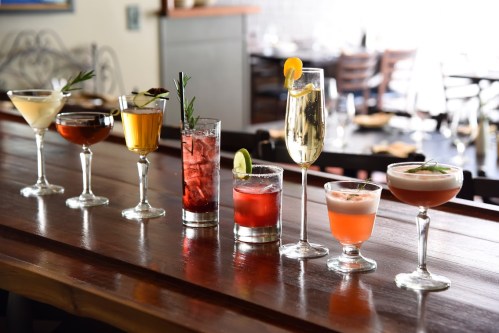 A variety of colorful cocktails arranged on a wooden bar, showcasing different glasses and garnishes.