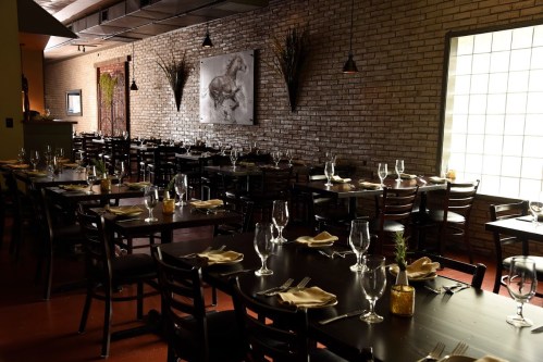 A dimly lit restaurant with wooden tables, chairs, and elegant table settings, featuring a large artwork on the wall.