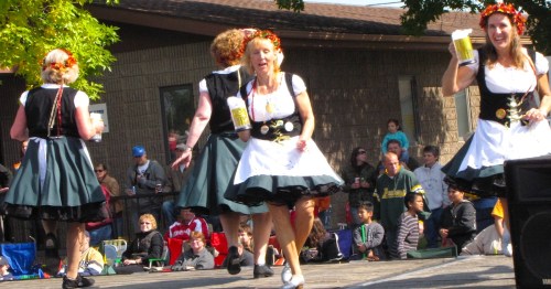 Women in dirndls dance on a stage while holding steins of beer.