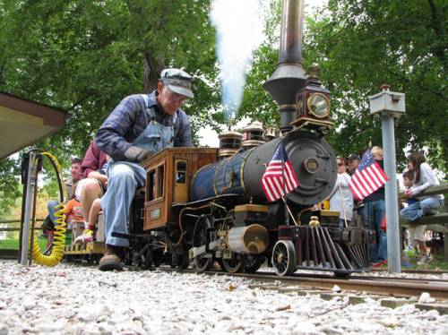 A man operates a miniature steam train adorned with small American flags, surrounded by spectators in a park.