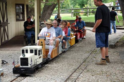 A miniature train with passengers rides along a gravel track, while a conductor stands nearby. People watch from the side.