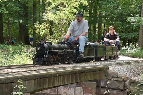 A miniature steam train travels along a track in a wooded area, with two people riding on it.