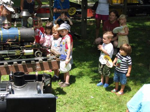 A group of children watching a miniature train at a park, some holding plates, with adults in the background.