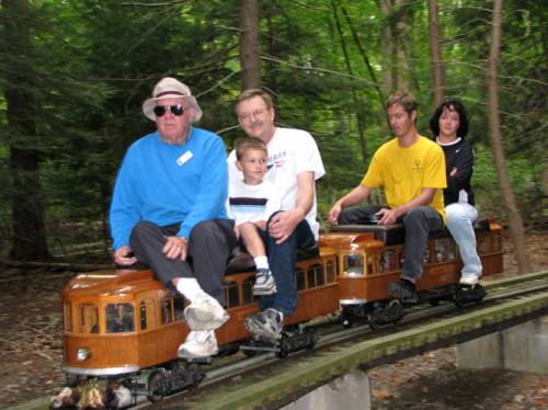 A group of four people riding a miniature train on a wooded track, enjoying a fun outdoor experience.