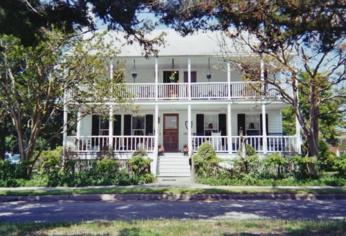 A large, two-story white house with a wraparound porch, surrounded by trees and greenery.