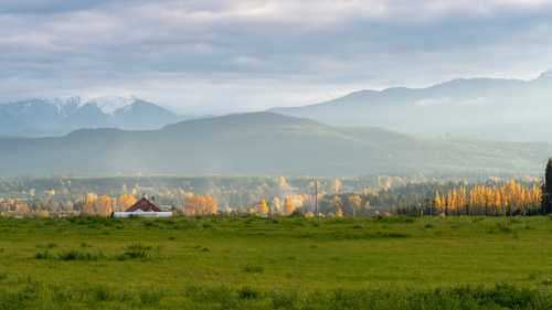 A serene landscape featuring a green field, a red barn, and mountains in the background with autumn foliage.