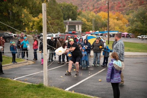A crowd watches as a man swings a slingshot to launch a pumpkin at a fall festival. Colorful trees in the background.