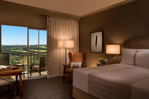 Cozy hotel room with a king bed, armchair, desk, and large window showcasing a scenic view of green hills.