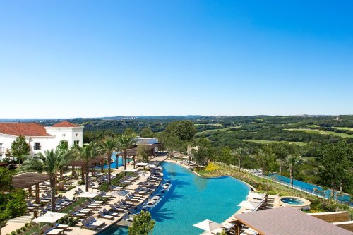A scenic view of a resort pool surrounded by palm trees and lounge chairs, with rolling hills in the background under a clear blue sky.