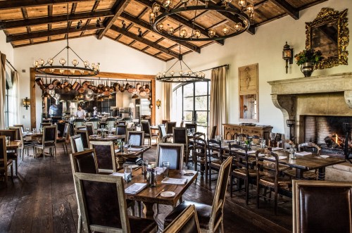 A spacious restaurant with wooden beams, elegant tables, and a cozy fireplace, featuring a visible kitchen area.