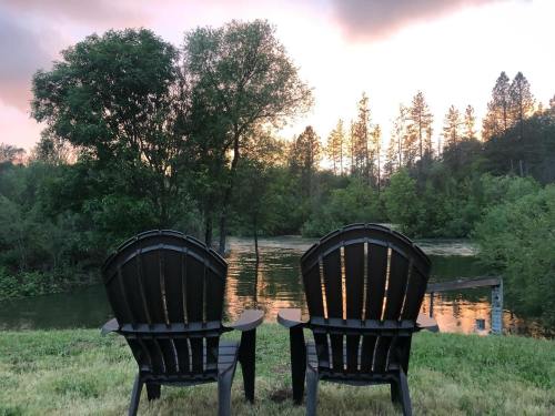 Two black Adirondack chairs facing a serene river at sunset, surrounded by trees and lush greenery.