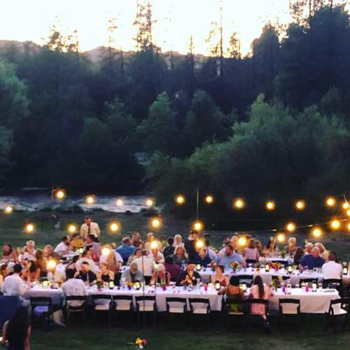 A large outdoor dinner party with guests seated at long tables, surrounded by trees and string lights at sunset.