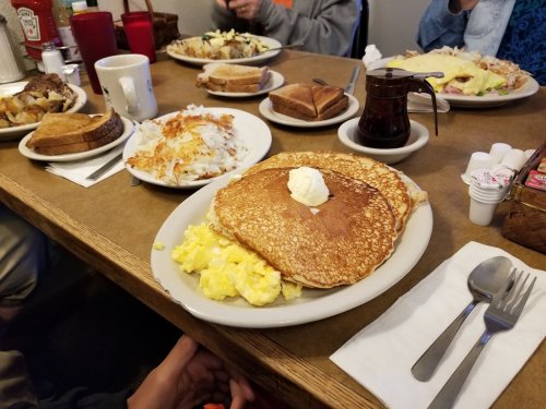 A table set with breakfast plates, including pancakes, scrambled eggs, toast, and hash browns, with coffee and syrup.