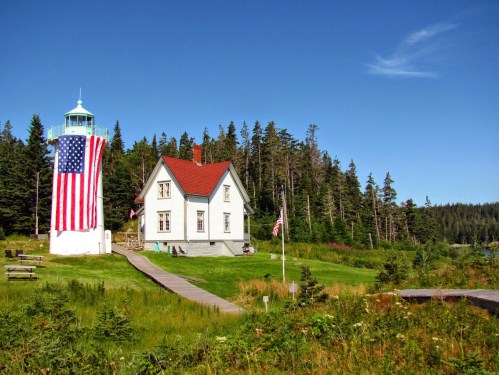 A lighthouse with an American flag draped on it, next to a white house surrounded by trees and green grass.