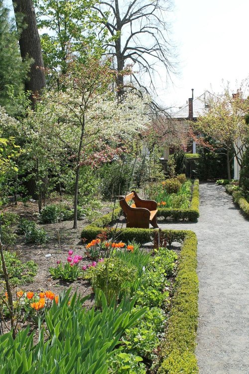 A serene garden path lined with colorful flowers, trees in bloom, and a rustic wheelbarrow amidst lush greenery.