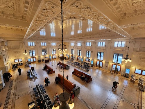 A spacious, ornate train station interior with high ceilings, decorative details, and waiting areas filled with people.
