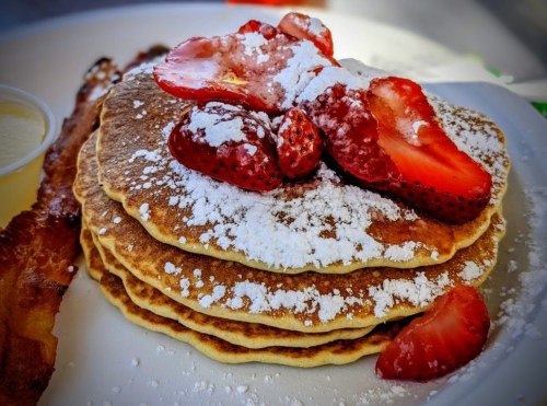 A stack of pancakes topped with sliced strawberries and powdered sugar, served with a side of bacon.