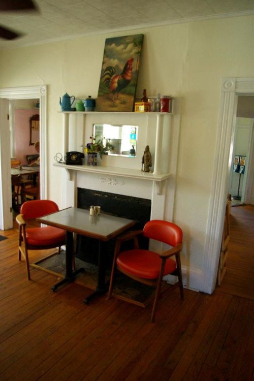 Cozy dining area with a wooden table, two orange chairs, and a decorative shelf above a fireplace.