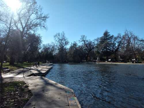 A serene park scene featuring a calm pond, trees, and a sunny sky, with people walking along the water's edge.