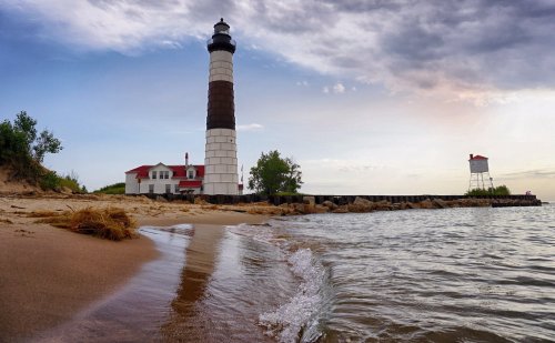 A lighthouse stands by the shore, with gentle waves lapping at the sandy beach under a cloudy sky.
