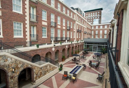 A courtyard with brick buildings, outdoor seating, and a fire pit, surrounded by greenery under a cloudy sky.
