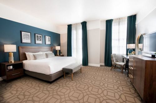 A modern hotel room featuring a king bed, stylish decor, a seating area, and large windows with curtains.