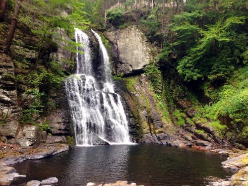 A serene waterfall cascading into a dark pool, surrounded by lush greenery and rocky terrain.