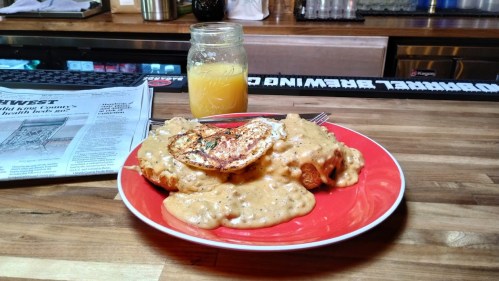 A red plate with biscuits and gravy topped with a fried egg, alongside a jar of orange juice and a newspaper.