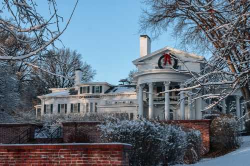 A snow-covered mansion adorned with a festive wreath, surrounded by trees and a brick wall in winter.