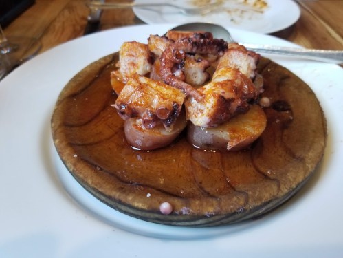Grilled octopus served on a wooden plate with sliced potatoes and a drizzle of sauce.