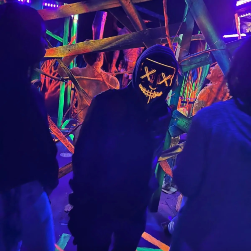 A person in a dark hoodie and a glowing mask stands among colorful, eerie decorations in a dimly lit space.