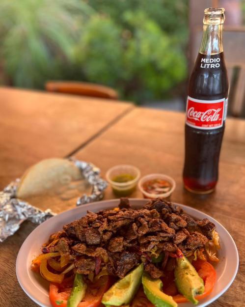 A plate of tacos topped with grilled meat, avocado, and peppers, alongside a bottle of Coca-Cola and sauces.