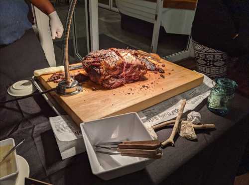 A large, roasted piece of meat on a wooden board, with utensils and a glass nearby, set on a dark table.