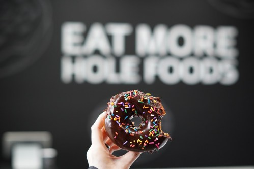 A hand holding a chocolate donut with colorful sprinkles in front of a "Eat More Hole Foods" sign.