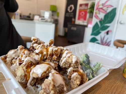 A takeout container filled with fried chicken drizzled with sauce, served over rice and accompanied by broccoli.