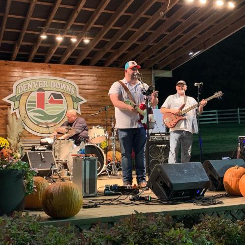 A band performs on stage at Denver Downs, with a guitarist, bassist, and drummer, surrounded by autumn decorations.