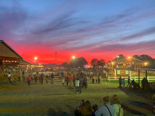 A vibrant sunset casts a colorful sky over a bustling fairground with people enjoying the evening.