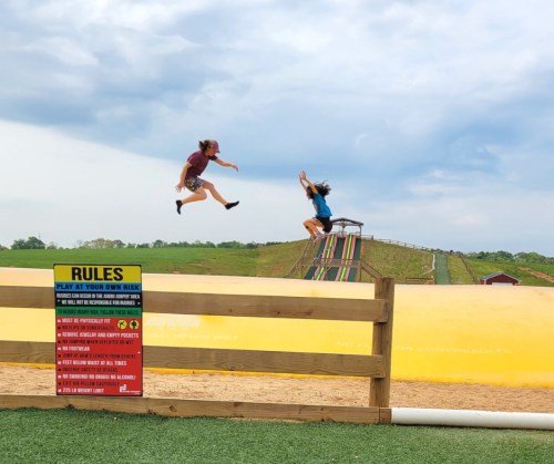 Two children jumping joyfully in the air near a large inflatable slide, with a rules sign in the foreground.