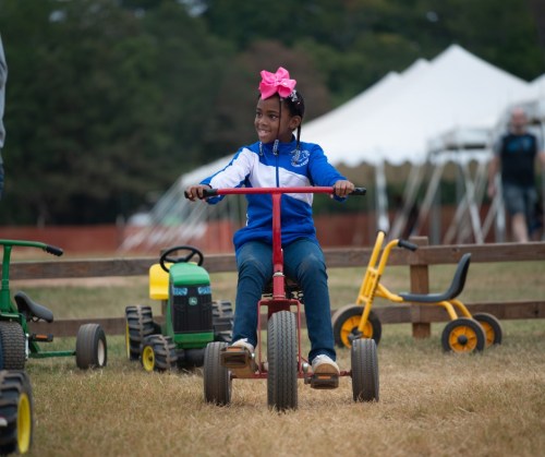 A young girl with a pink bow smiles while riding a red tricycle in a grassy area with toy tractors nearby.