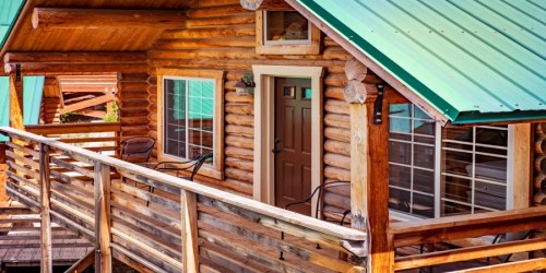 A rustic log cabin with a green metal roof, featuring a wooden deck and chairs on the porch.