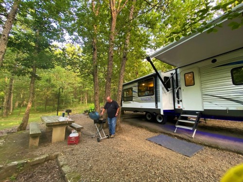 A man grills outside a camper in a wooded area, with a picnic table nearby and trees surrounding the scene.