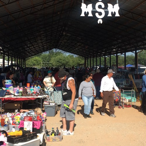 A bustling outdoor market with vendors and shoppers browsing various items under a large roof.