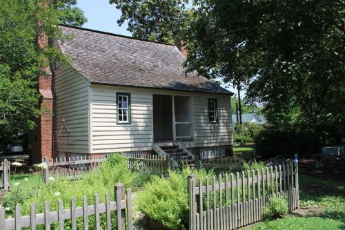 A small, historic house with a gabled roof, surrounded by greenery and a wooden fence.
