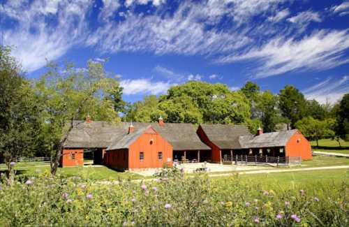 A picturesque red barn surrounded by green trees and a blue sky with fluffy clouds, set in a rural landscape.